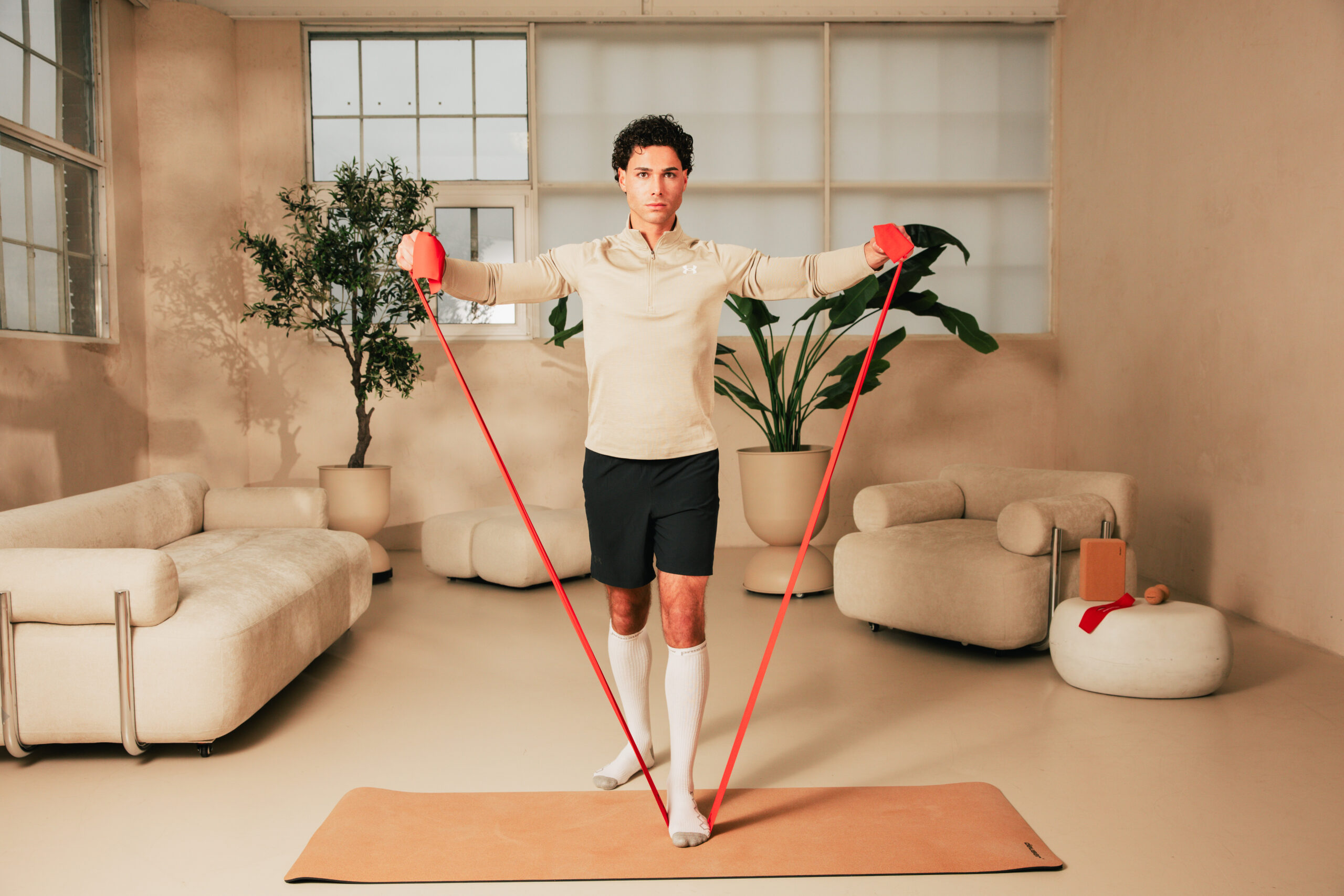 251122_DEUSER-129 - Deuser Sports | Solingen Man performing a standing balance exercise with red resistance bands indoors, arms extended to the sides on a yoga mat.