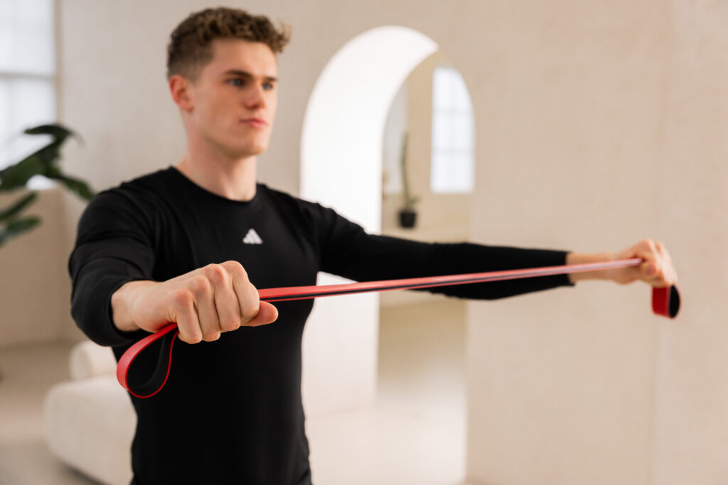 Man in a black shirt stretches a red resistance band with both hands indoors, exercising his arms in a living room setting.