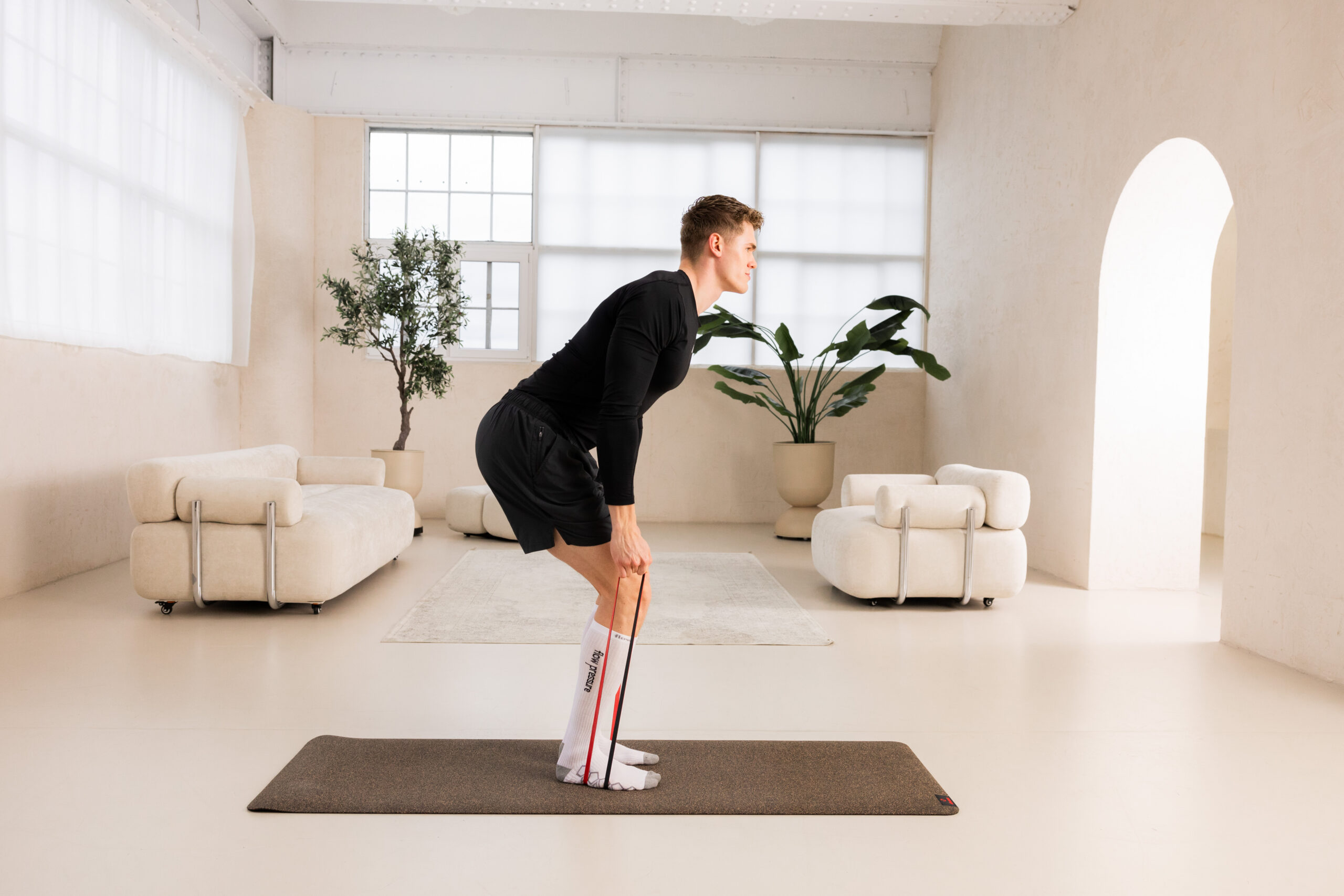 251122_DEUSER-2-3 - Deuser Sports | Solingen Man in black athletic wear bent over on a mat, using a resistance band for a hip hinge exercise in a bright, minimalist living room with plants and white seating.
