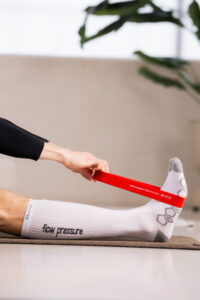 251122_DEUSER-38 - Deuser Sports | Solingen Close-up of a person performing a leg stretch with a red resistance band looped around the ankle, wearing white socks on a mat.