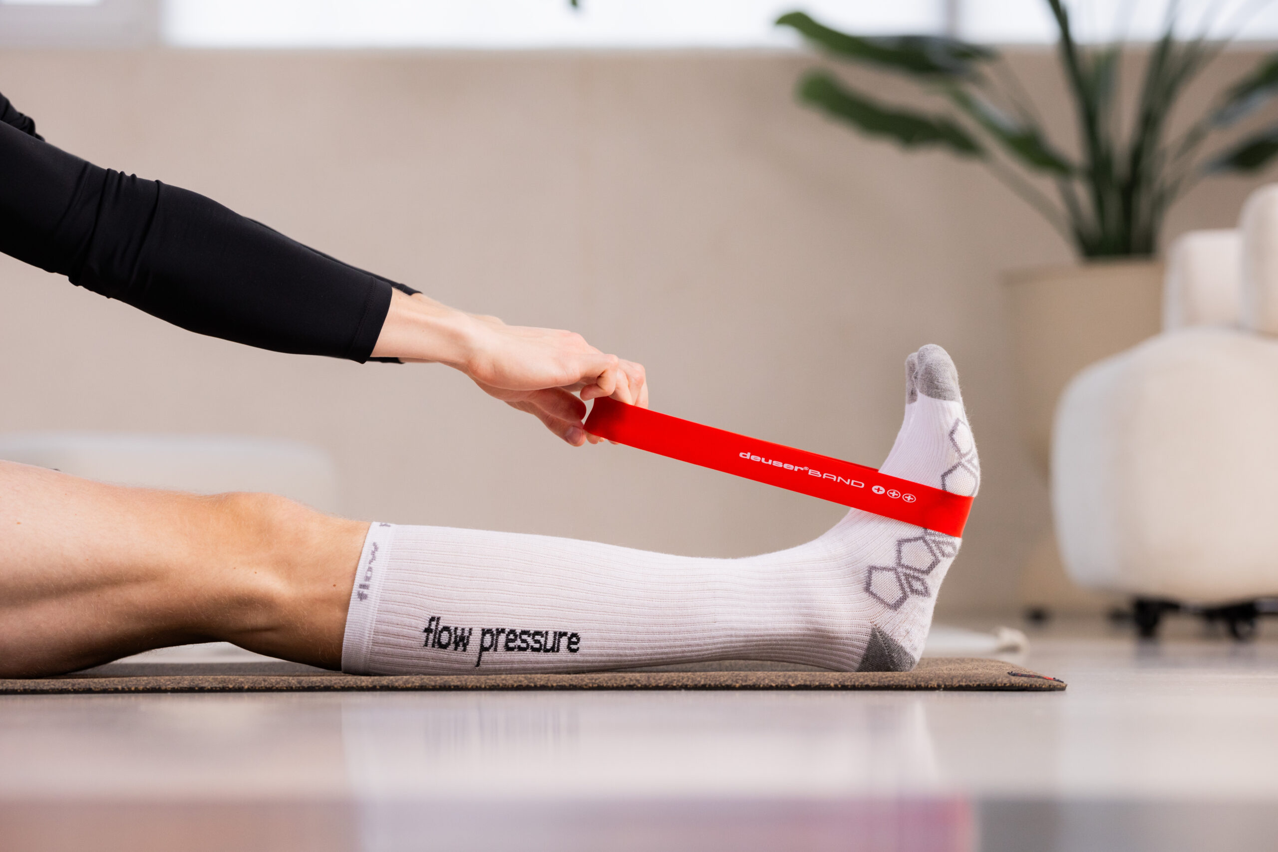 251122_DEUSER-40 - Deuser Sports | Solingen Person's foot and lower leg on a mat with a red ankle resistance band being held by another person during a leg exercise outside a living room setting.