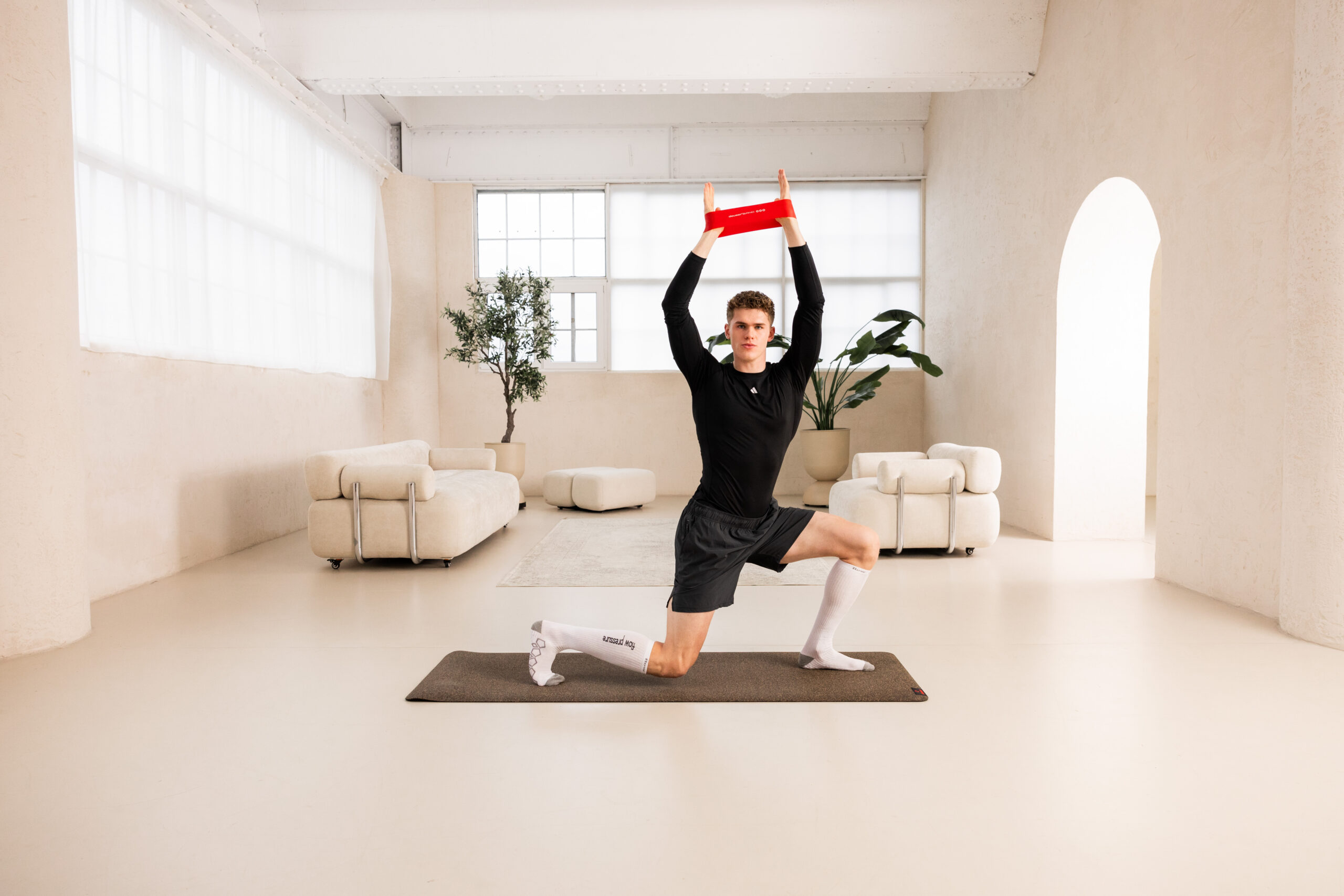 251122_DEUSER-5 - Deuser Sports | Solingen Young man in black athletic wear on a yoga mat raises a red resistance band overhead in a bright minimalist living room.