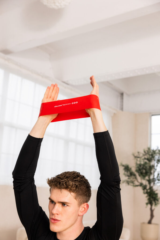 Young man in a black long-sleeve shirt raises a red resistance band overhead during a workout in a bright studio.