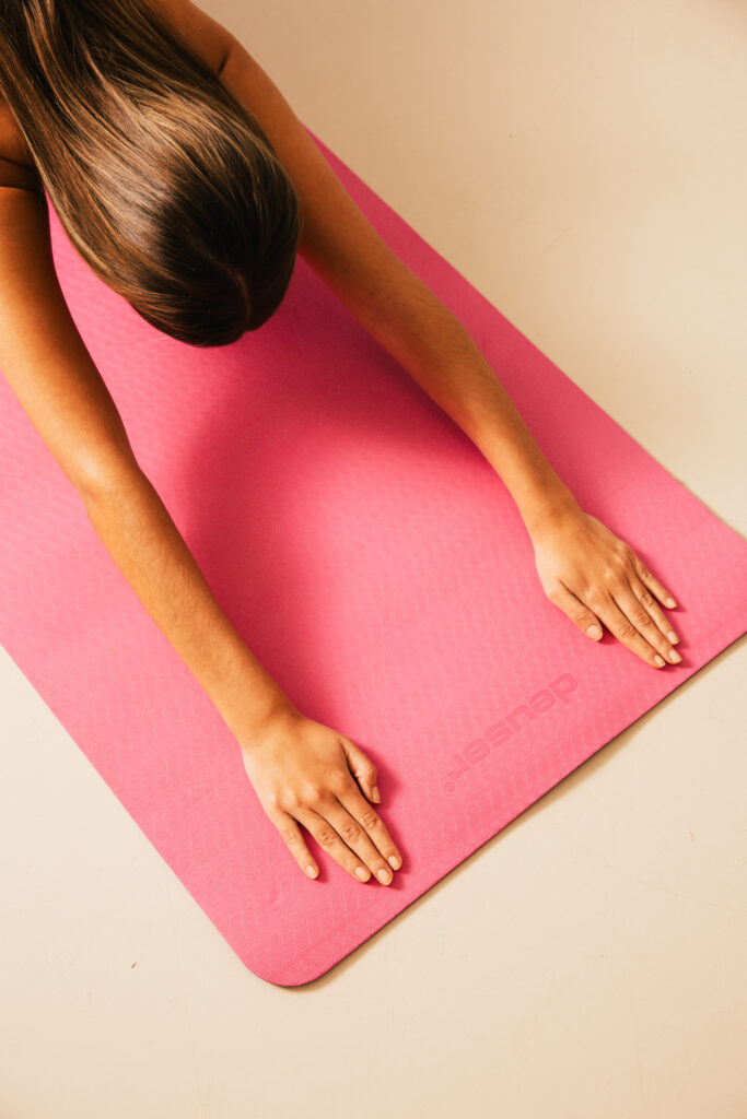 Top-down view of a person with long hair, arms extended on a pink yoga mat, mid-stretch.