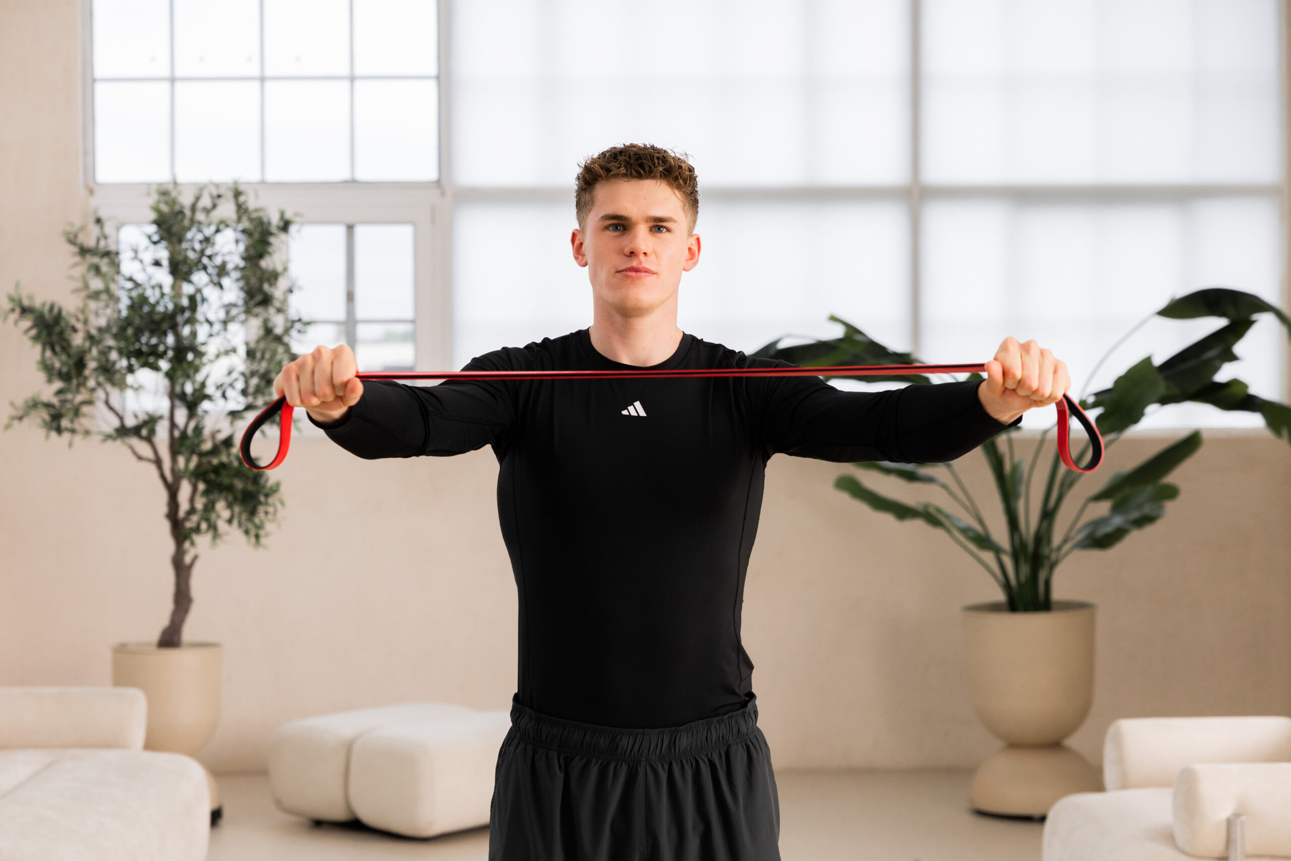 251122_DEUSER-13 - Deuser Sports | Solingen Young man in black athletic wear extends a red resistance band in a bright room with plants in the background.