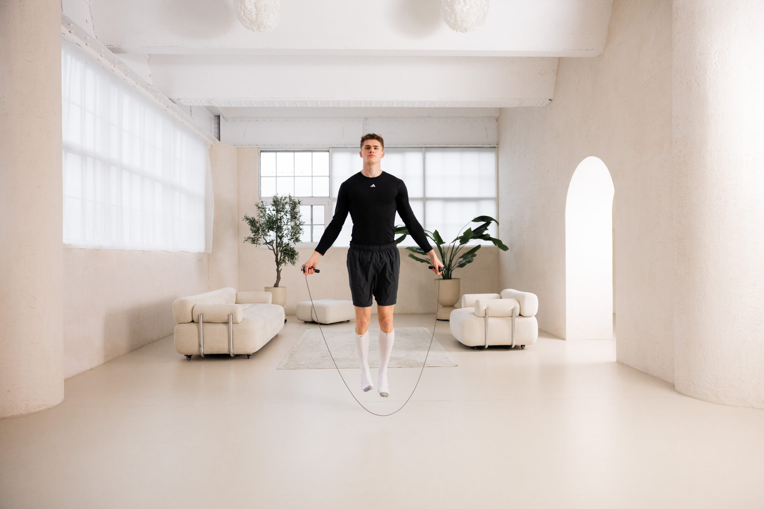 251122_DEUSER-18 - Deuser Sports | Solingen Man in black athletic top and shorts jumping rope in a bright minimalist living room with plants and cream furniture.