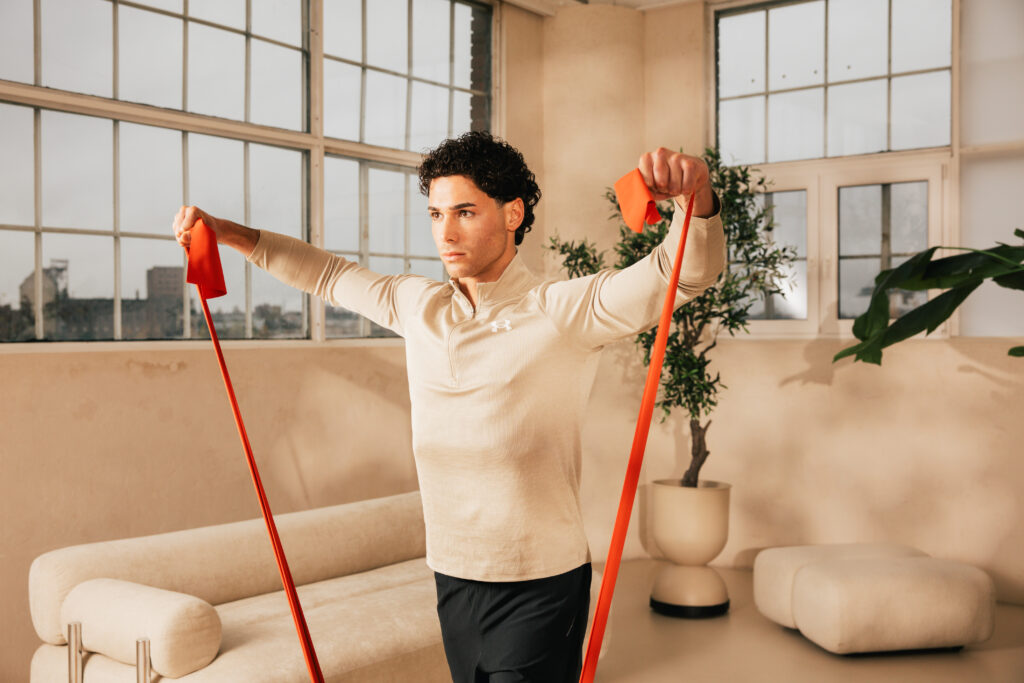 Man wearing a beige athletic top uses red resistance bands in a bright living room for an upper-body workout with arms extended.