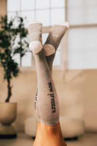251122_DEUSER-95 - Deuser Sports | Solingen Two legs raised in the air, wearing beige knee‑high socks with a pattern, in a bright indoor space with a plant in the background.