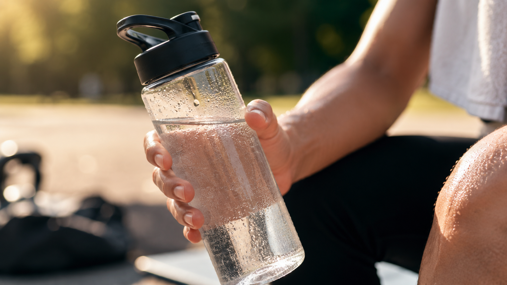 efe9743e-0592-48e1-9976-a3482edb98ee - Deuser Sports | Solingen Close-up of a person’s hand gripping a clear plastic water bottle with condensation, outdoors in warm sunlight.